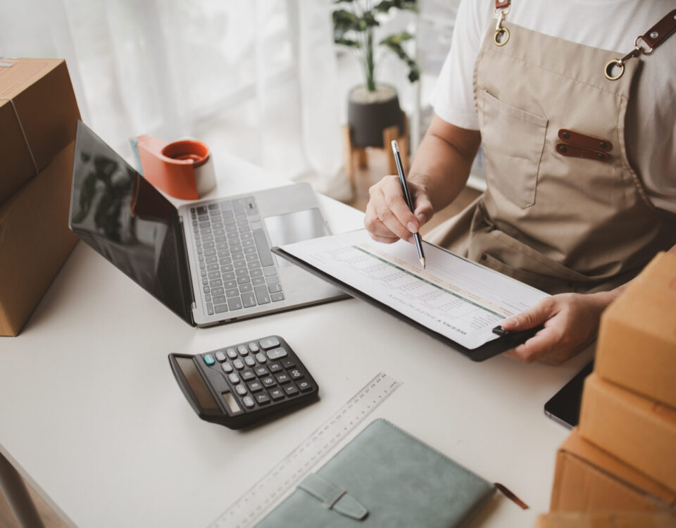 Business owner in an apron reviewing a checklist beside a laptop and parcels before moving stock into a Sunninghill storage unit